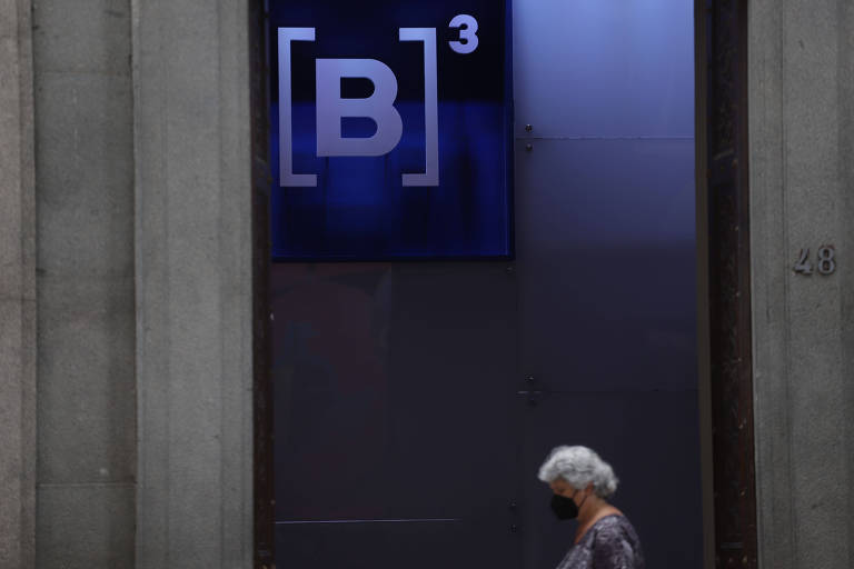 Mulher de cabelos brancos e máscara preta passa na frente da sede da B3, Bolsa de Valores de São Paulo; ao fundo, aparece a logo da B3, que tem fundo azul e letras brancas