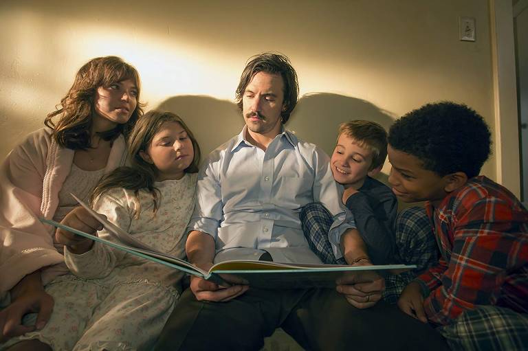 Um homem e uma mulher brancos de cerca de 30 anos estão sentados, de pijama, junto com três crianças, duas brancas e uma negra. Todos leem um livro