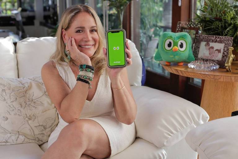 Susana Vieira está sentada num sofá, sorri e mostra um celular para a câmera