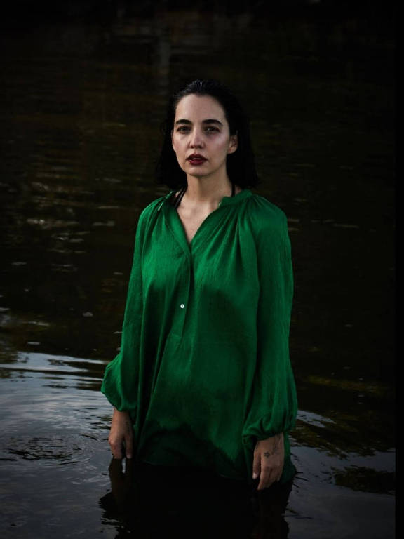 A artista Rits Wainer está dentro de uma lagoa, com água até as pernas; ela é branca, tem cabelos curtos e pretos e veste uma bata verde