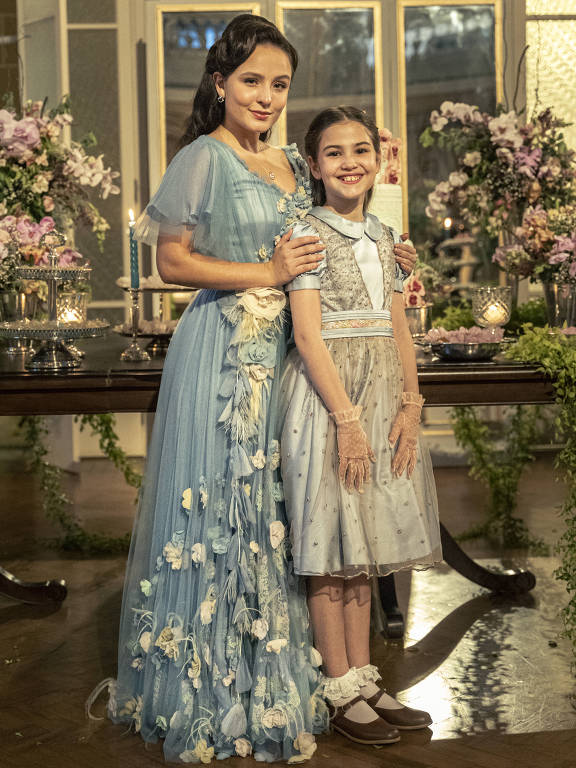 As atrizes Larissa Manoela e Sofia Budke estão lado a lado, em pé, com vestidos de festa em um salão de baile