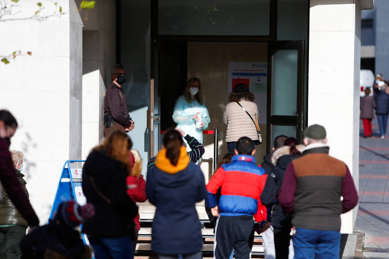 Pessoas em frente a um centro médico; na porta do centro, uma profissional de saúde com roupas de proteção
