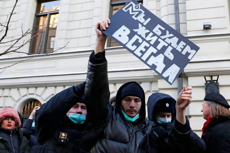 Homem de jaqueta preta levanta cartaz escrito em russo enquanto é capturado por policiais