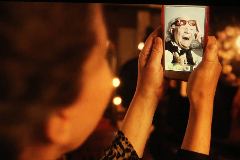 Mãos femininas seguram celular com a tela voltada para a lente. Em quadro o ator Ney Latorraca com camisa social e óculos escuros aparece em close divertido. Alto astral.