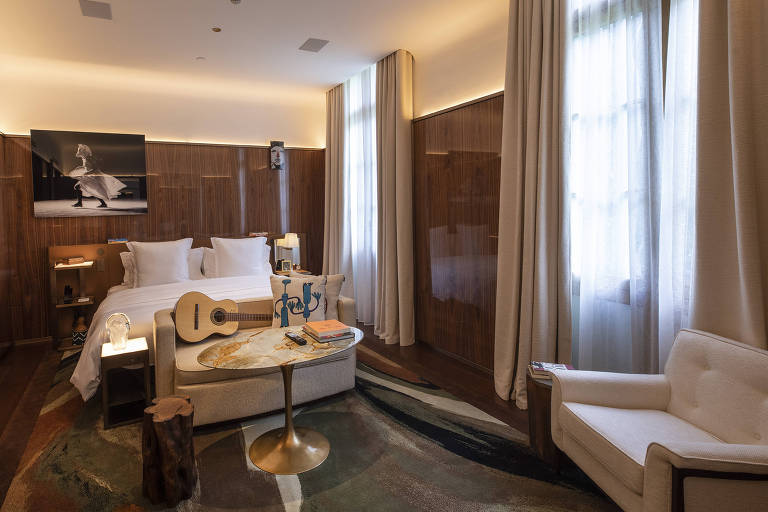 Quarto de hotel com revestimento amadeirado nas paredes, com uma cama de casal com lençóis claros, com um violão aos pés da cama.