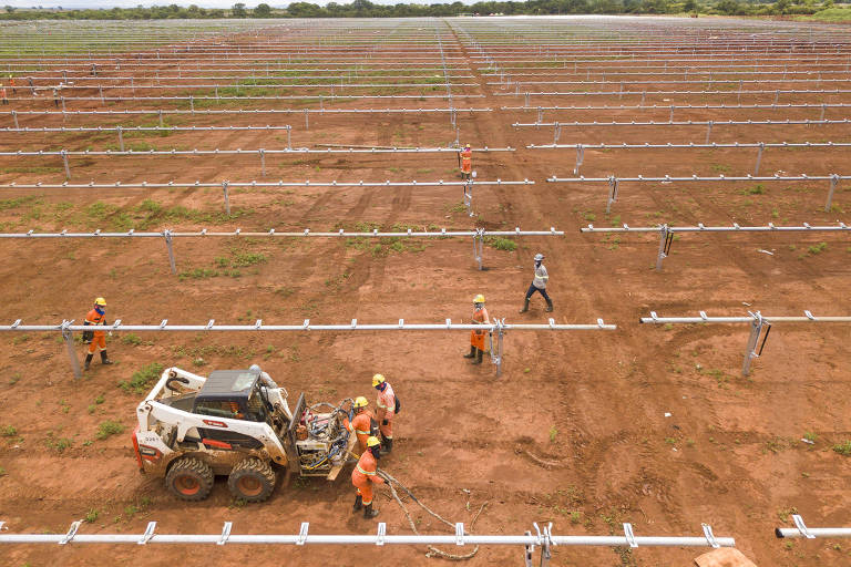 Vista aérea mostra um campo de terra com trabalhadores que vestem laranja