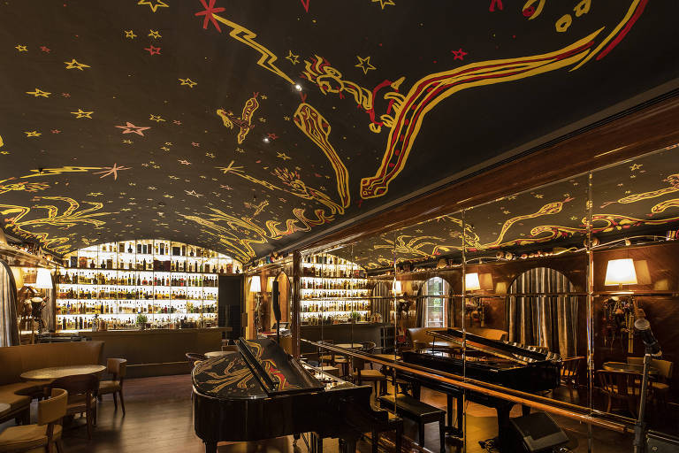 salão de restaurante com piano ao centro, teto azul escuro adornado com detalhes dourados; parede com garrafas ao fundo