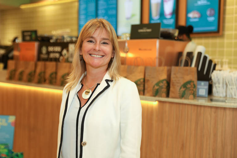 Claudia Malaguerra, diretora geral da Starbucks no Brasil, aparece sorrindo, dentro de uma loja. Ela é uma mulher branca com cabelos loiros e sorri para a câmera