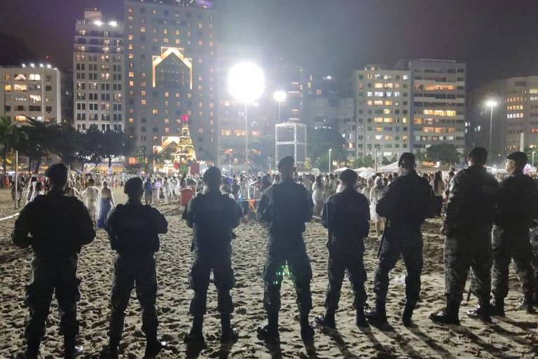 Policiais aparecem enfileirados em praia, durante a noite, em frente a prédios iluminados