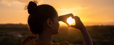 Silhueta contra-luz de mulher, com coque, fazendo sinal de coração com as mãos em direção ao pôr-do-sol