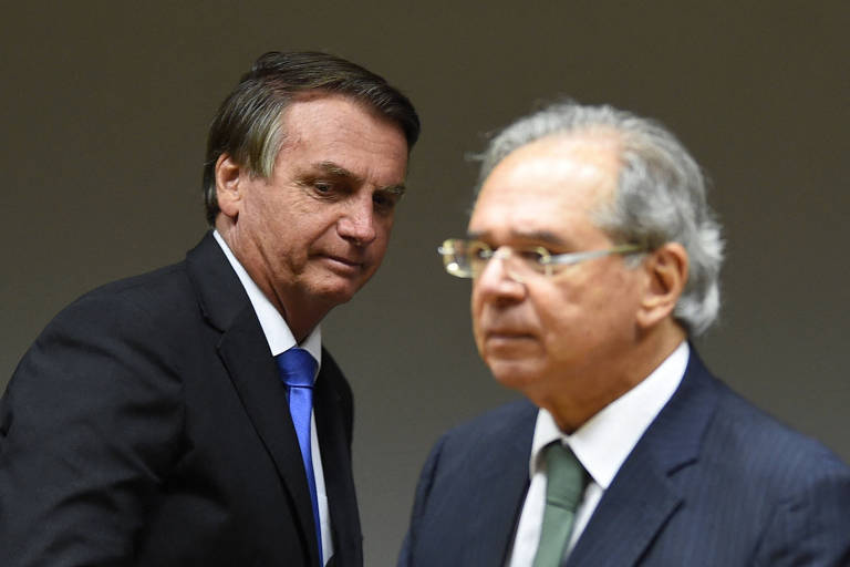 O presidente Jair Bolsonaro, de pé, passa ao lado do ministro Paulo Guedes