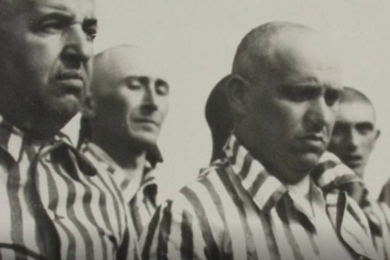 Foto em preto e branco mostra homens com cabeça raspada, usando um uniforme listrado; eles são prisioneiros de um campo de extermínio nazista