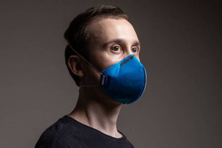 Homem de camisa preta em fundo preto usa uma mascara azul do tipo N95 contra o coronavírus