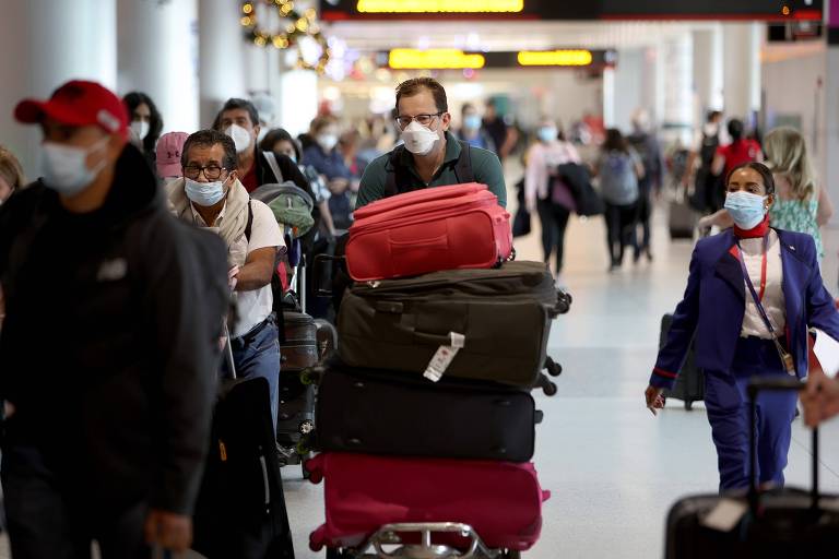 Homem usando máscara carrega carrinho com malas em corredor de aeroporto, em que estão outros passageiros carregando malas e usando máscaras 