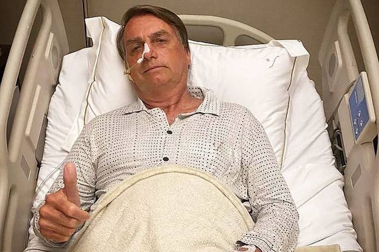 O presidente Bolsonaro está deitado numa cama de hospital, com pijama claro e um acesso no nariz