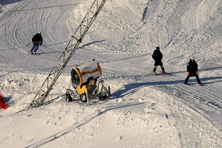 Homens andam em montanha de neve, ao lado de uma máquina