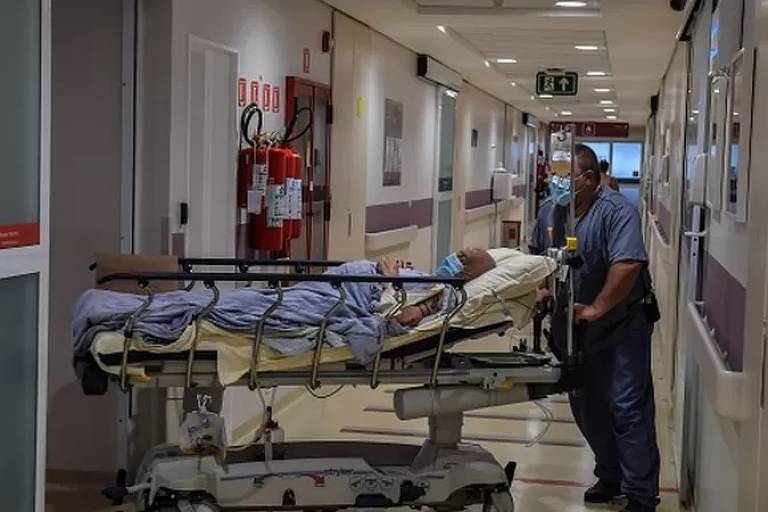 Imagem em primeiro plano mostra paciente deitado em uma cama de UTI sendo empurrado por um profissional da saúde