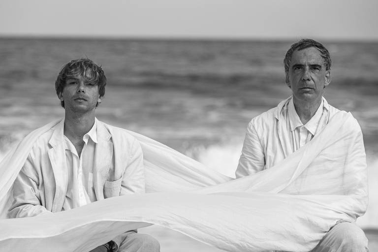 Dois homens sentados de costas para o mar são transpassados por longos panos brancos