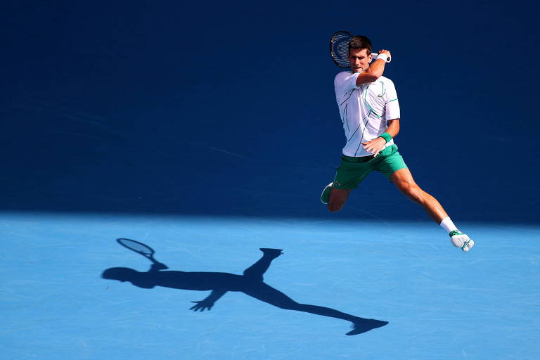 Djokovic saltando e rebatendo uma bola no fundo de quadra, com o registro de sua sombra na quadra