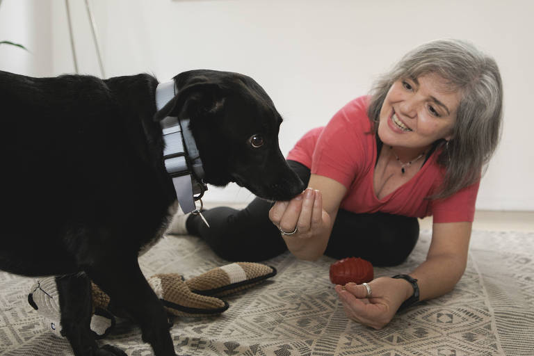 Sentada no chão, a pet sitter Mirna Moura dá ração na boca de um cachorro preto de porte médio