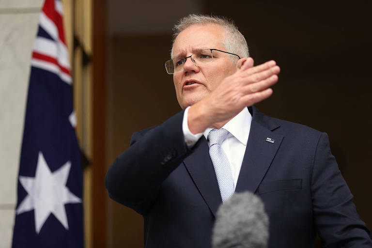 Homem de terno faz movimento com o braço direito durante discurso em frente a bandeira da Austrália