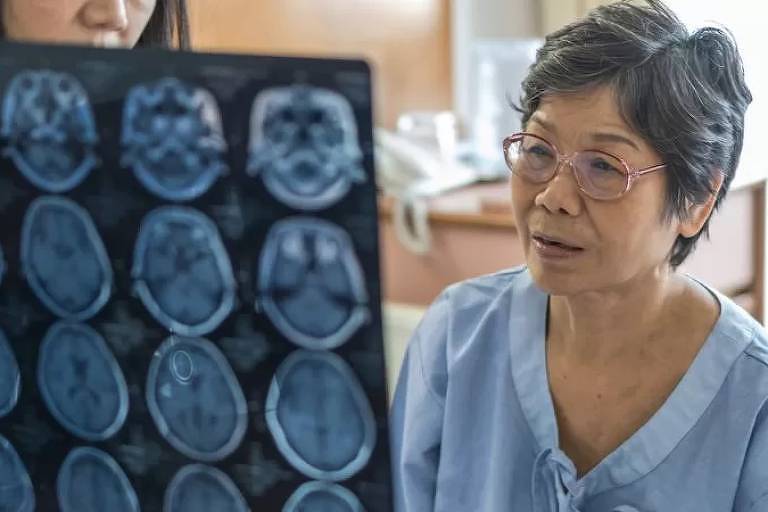 Imagem em primeiro plano mostra mulher grisalha de óculos olhando para um resultado de raio x, que está sendo segurado por outra pessoa.