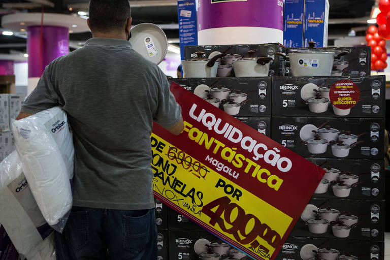 Cliente é visto de costas, segurando dois travesseiros e observando promoção de panelas