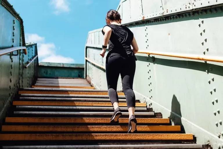 Imagem em primeiro plano mostra mulher com roupa de ginástica de costas subindo uma escada