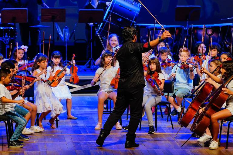 Emfoto colorida um grupo de crianças tocam instrumentos de cordas (violoncelo, viola e violino), regidas por um maestro, em um palco de um teatro.
