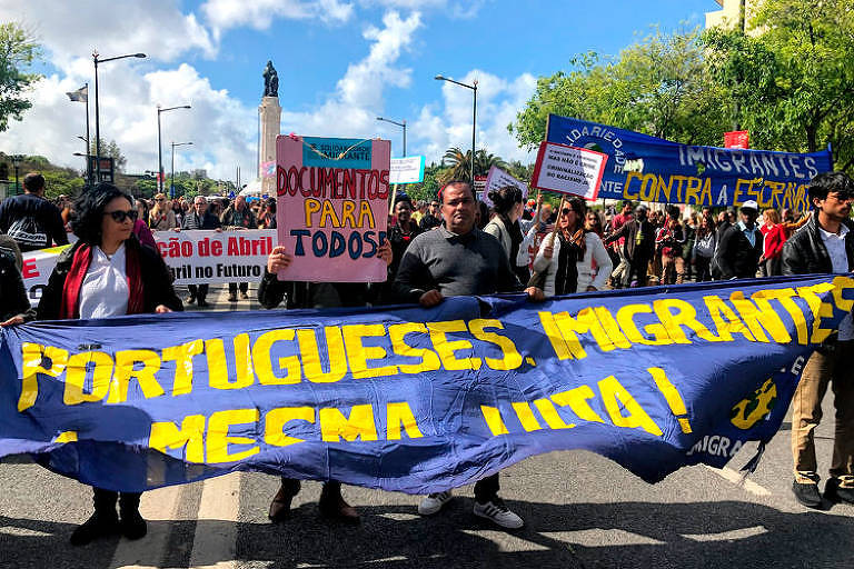 Imigrantes  protestam por seus direitos durante desfile do 25 de abril em Lisboa. Imagem mostra diversas pessoas segurando diferentes cartazes. No maior deles está escrito "Portugueses e imigrantes, mesma luta". Em outro, mais ao fundo, há a inscrição "documentos para todos"
