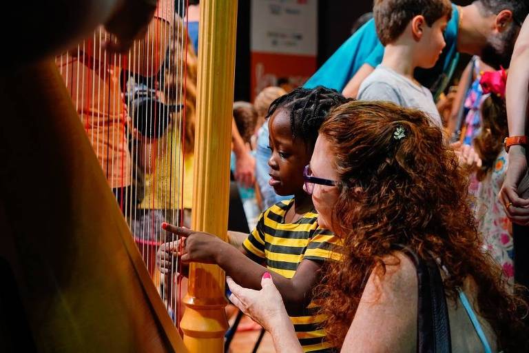 Em foto colorida, uma criança negra é auxiliada pelas mãos de uma professora branca a tocar uma harpa.