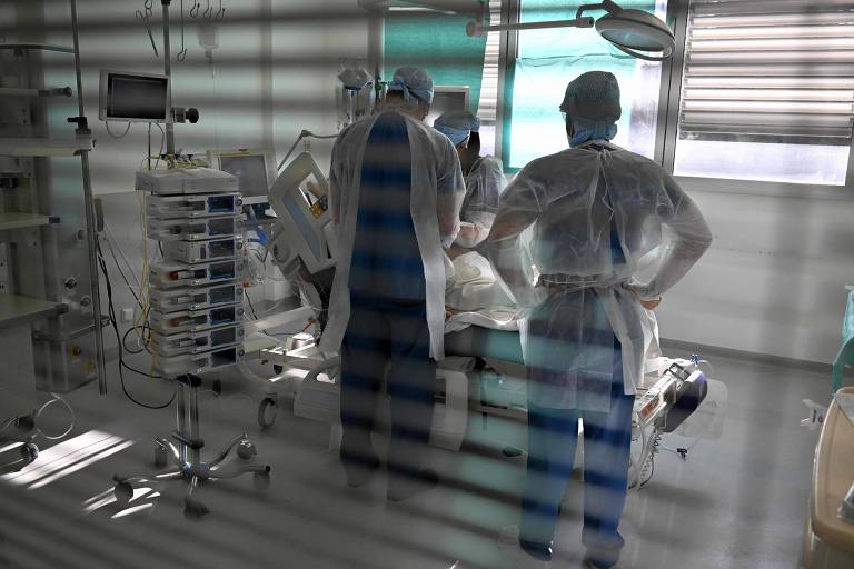 Vistos através de uma janela com persiana entreaberta, médicos olham paciente que está deitado em leito