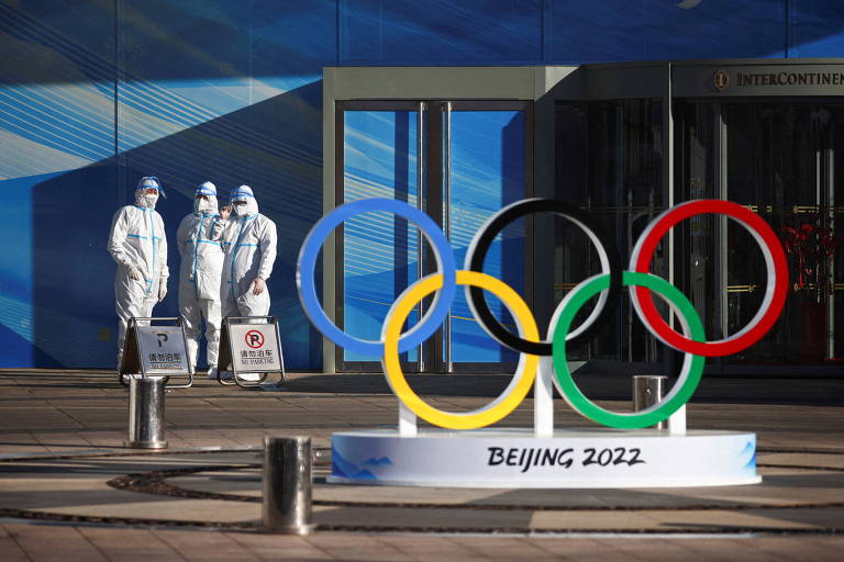 Ao fundo, três pessoas com roupas especiais brancas estão de pé. As roupas cobrem os corpos inteiros. Em primeiro plano há os anéis olímpicos sobre um suporte em que está escrito "Pequim 2022"