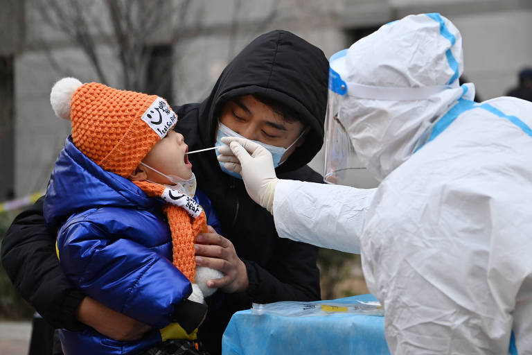 Homem segura criança enquanto pessoa em equipamento de proteção insere cotonete na boca da criança