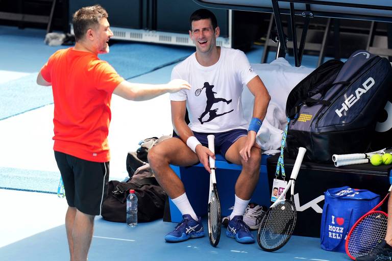 Djokovic sentado e sorrindo em conversa com outro homem, que está de pé na sua frente