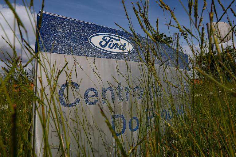Imagem aberta, feita com grande angular, mostra em primeiro plano mato na frente de placa da fábrica da Ford em Camaçari