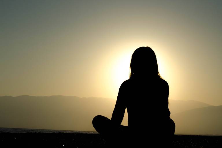 Silhueta de uma mulher, à sombra, com sol ao fundo, meditando