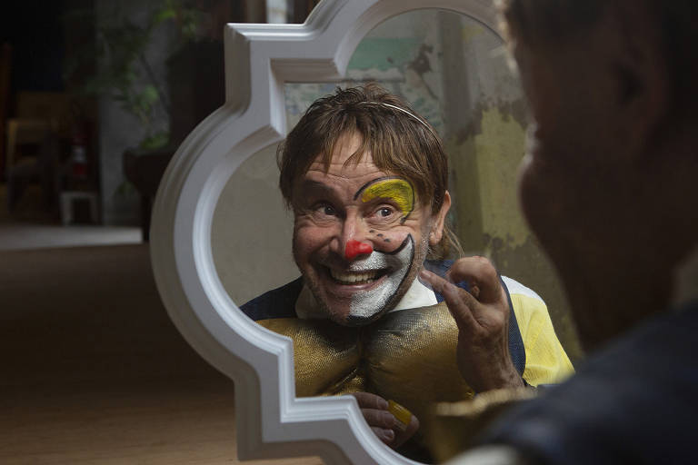 Homem sorridente, com maquiagem e nariz de palhaço, olha para espelho