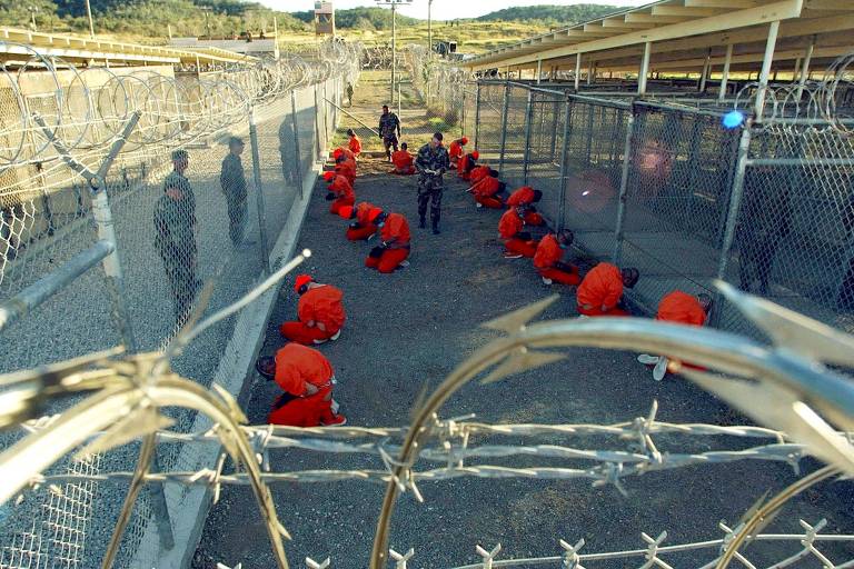 Detidos em macacões laranja sentam em uma área de detenção, enquanto são observados pelos militares dos EUA, no acampamento temporário de X-Ray, que mais tarde foi fechado e substituído pelo Acampamento Delta, dentro da base naval da Baía de Guantánamo