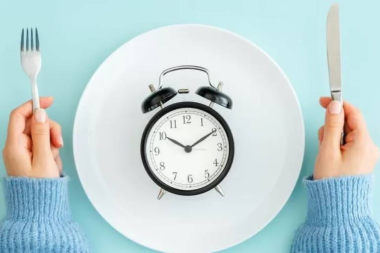 Imagem aérea e em close mostra as mãos de uma pessoa segurando talheres. No meio, há um relógio do tipo despertador dentro do prato de refeição.