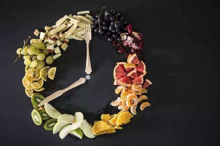 Pedaços de frutas formam um círculo e talheres são dispostos ao centro. A imagem remete a um relógio.