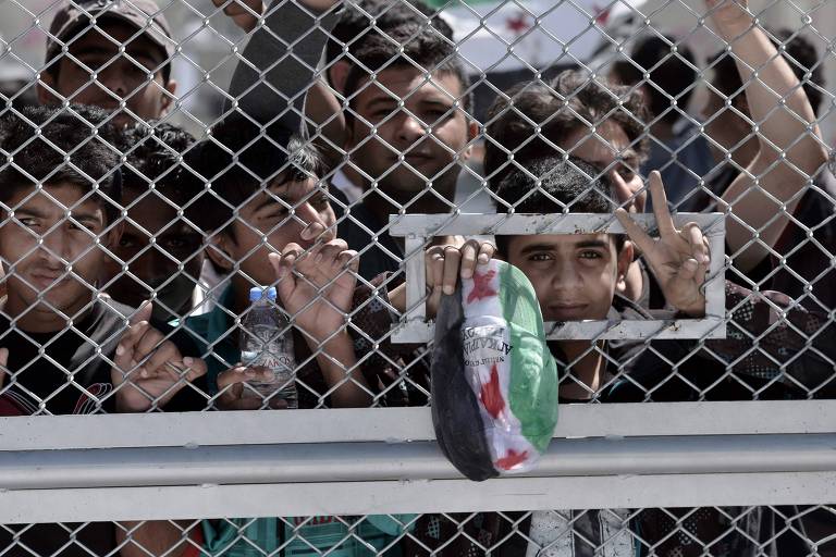vários jovens imigrantes sírios estão atrás de grades. em uma pequena abertura, um jovem faz um sinal com os dois dedos e mostra um boné nas cores branca, verde e preta da bandeira da Síria 