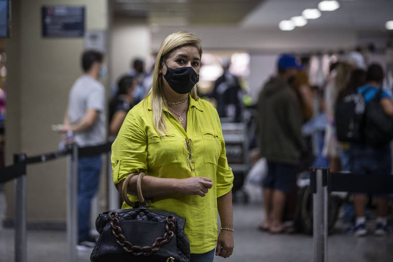 Mulher de amarelo e máscara preta, segurando uma bolsa preta, em frente a uma multidão no aeroporto