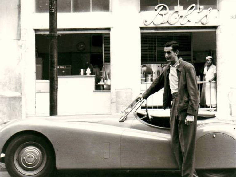 Foto preto e branca de homem em pé ao lado de carro antigo; no prédio atrás há um letreiro com o nome Bob's