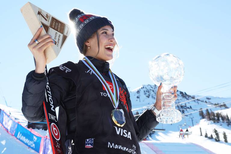 Esquiadora chinesa com roupas pesadas e touca sorri com dois troféus nas mãos