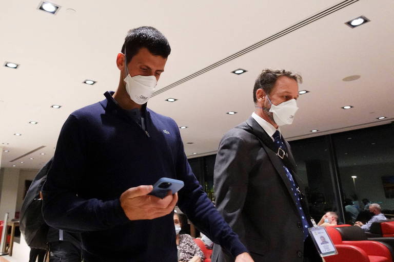 Djokovic anda olhando para o celular no aeroporto ao lado de um homem de terno; ambos estão de máscara