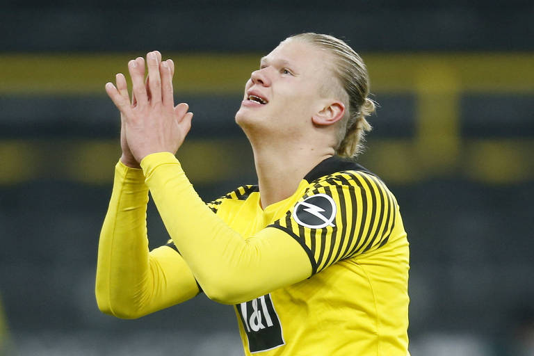 O norueguês Erling Haaland, avaliado pelo Transfermarkt em 150 milhões de euros, em jogo do do Borussia Dortmund; sua camisa é amarela, e ele está com as duas mãos abertas, juntas, enquanto olha para cima como se pedisse algo aos céus 