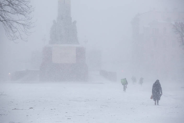 Em uma praça coberta de neve, algumas pessoas andam pela nevasca