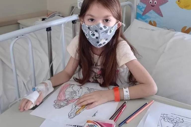 Imagem em primeiro plano mostra criança de máscara sentada em uma maca de hospital. Elas está com os braços apoiados sobre uma mesa com folhas de papel desenhadas e lápis de cor