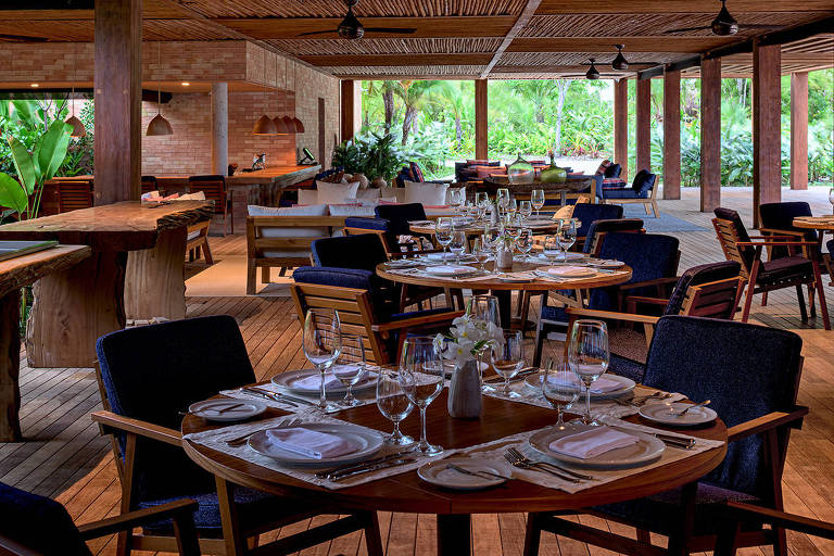 A foto mostra um salão de restaurante com mesas, onde predomina a madeira. As paredes de vidro mostram uma densa vegetação ao redor